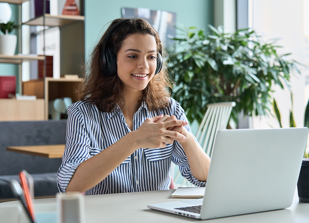 A smiling young woman wearing headphones and a striped shirt looking at a laptop while taking an online class